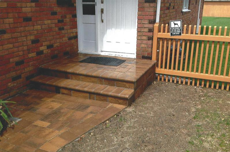 Brick steps lead to a white door. A brown brick path and a wooden fence border a small yard.
