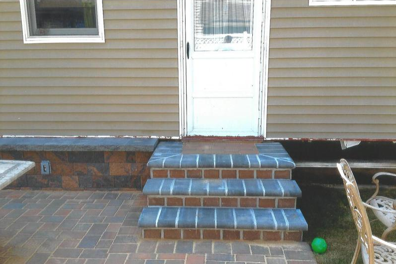 Brick steps leading up to a white door on a house with brown siding. Brick patio in front.