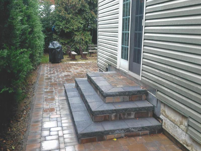 Brick steps leading to a door on the side of a house, with a paved walkway beside shrubs.