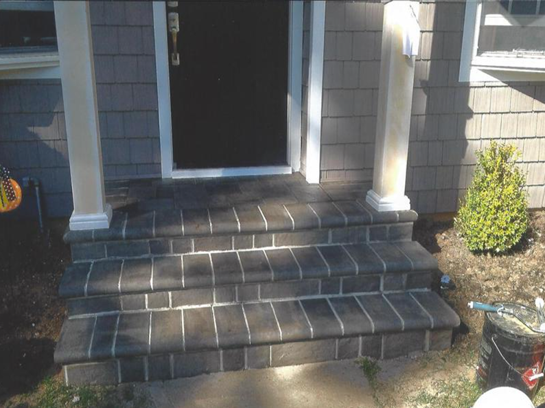 Brick steps lead to a front door with two white columns. Gray siding and a green bush are also visible.
