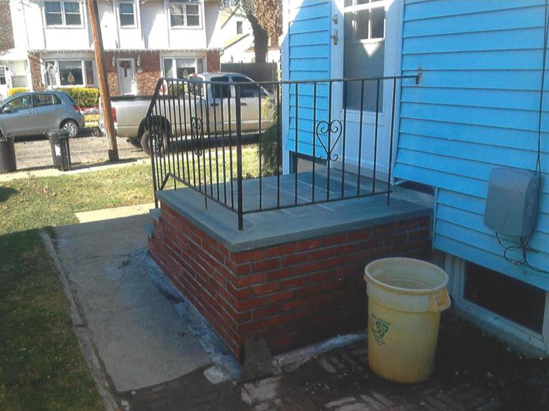 Brick steps leading to a blue house with a black railing. A yellow trash can sits next to the steps.