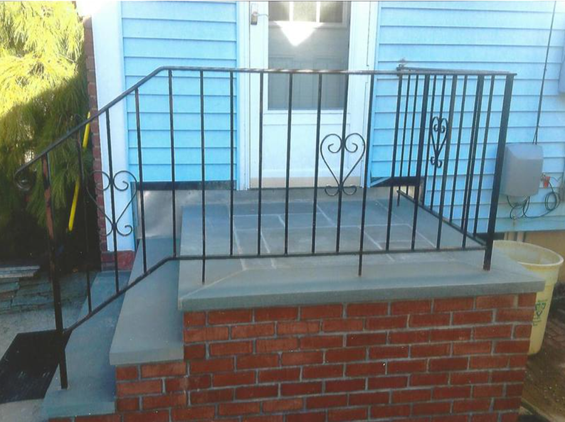 Black wrought iron railing on a brick and concrete porch in front of a blue house with a white door.
