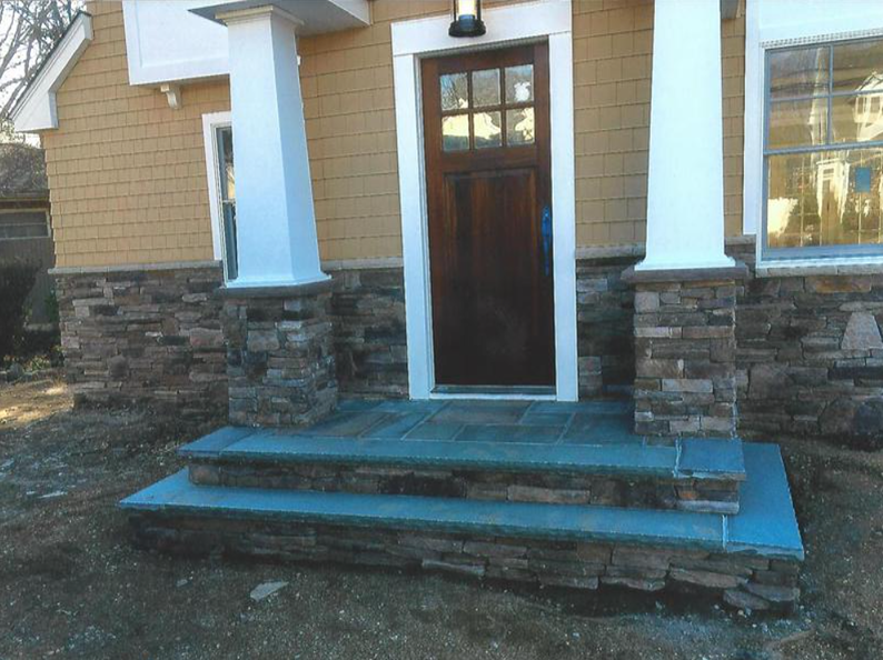 Stone steps lead to a wooden door under a porch with stone veneer columns.