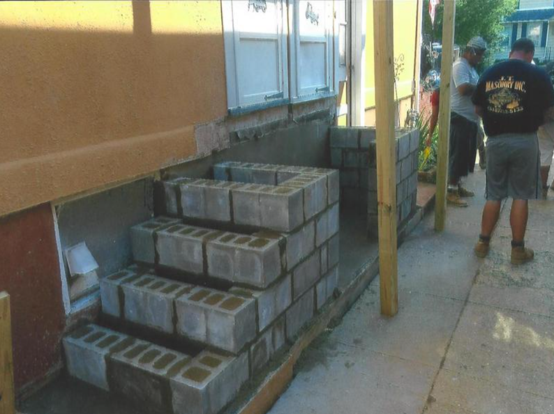 Workers building concrete steps next to a house with beige siding and white window shutters.
