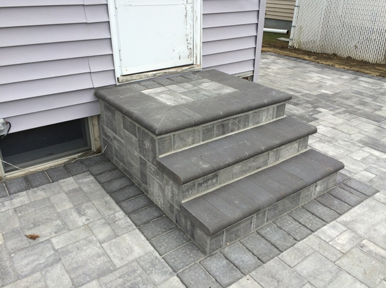 Three-step brick stairs leading to a white door on a house with a brick patio.