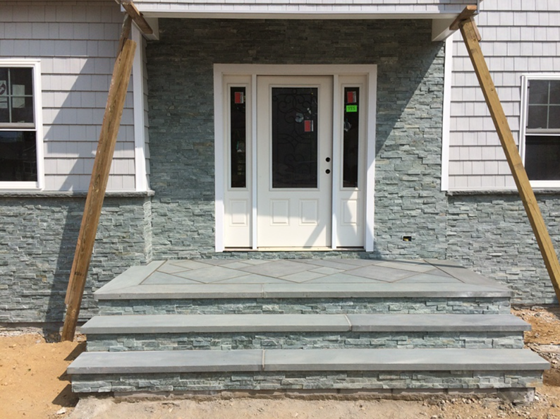 Stone steps lead to a front door with stone accents. The house has gray siding and two windows.