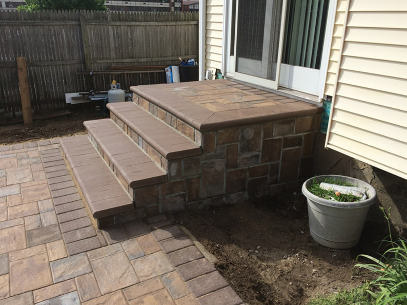 Stone steps lead to a patio door on a house, with a stone facade and a brown paver patio.