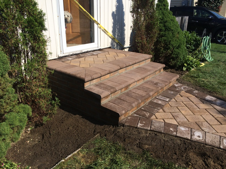 Brick steps leading to a house door with a pathway. Brown bricks, green lawn, and plants.