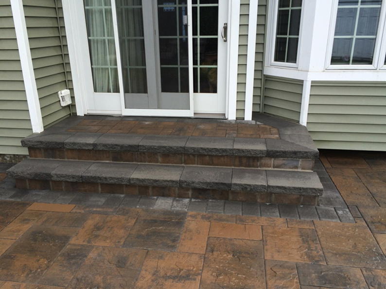 Brick patio with two steps leading to a sliding glass door on a house with green siding.