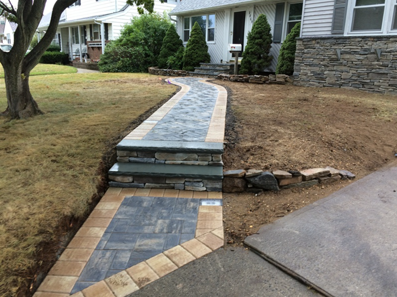 Stone walkway with steps leading to a house with tan and gray pavers, bordered by tan bricks.