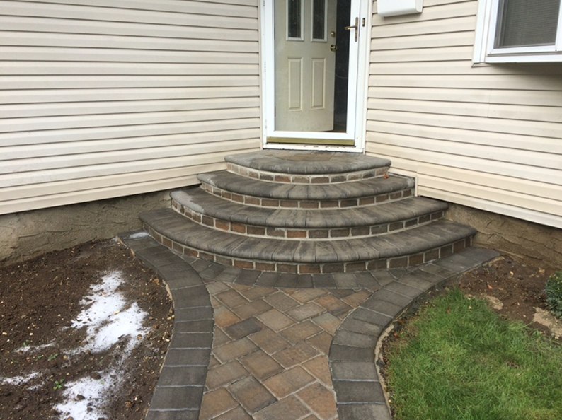 Brick steps leading to a beige door, with a paved walkway. Grass and dirt flank the stairs.