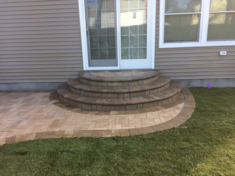 Brick patio with curved steps leading to a sliding glass door; tan siding and green grass.