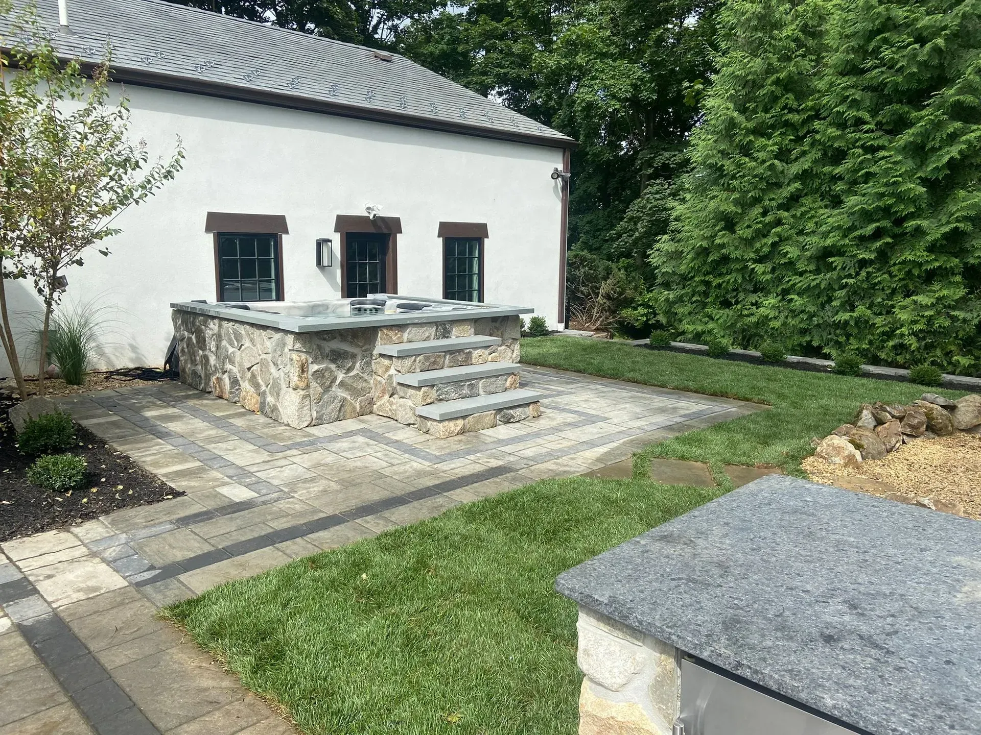 Hot tub with stone surround and steps, surrounded by pavers and lawn, beside a white building.
