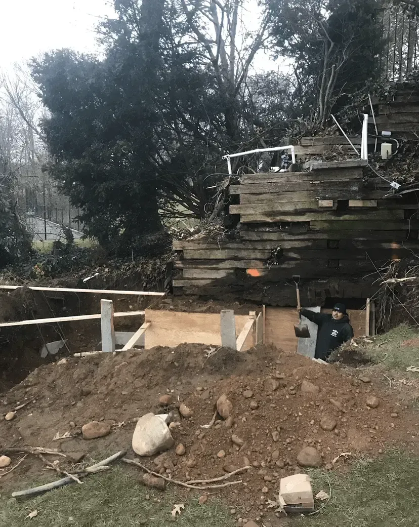 Construction site: worker digging in a trench next to a wooden retaining wall and pile of lumber.