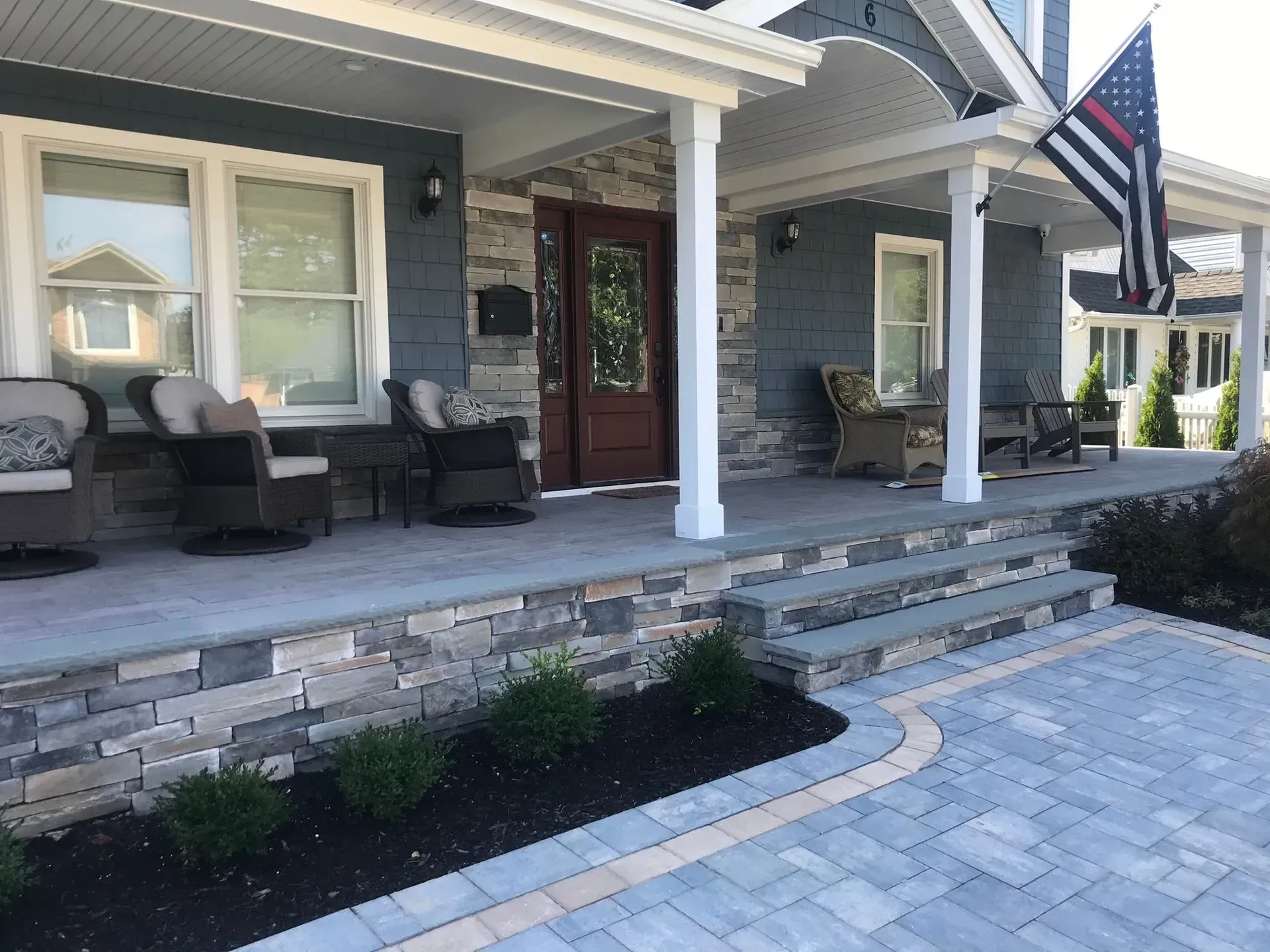 Front porch with stone facade, steps, and a brick driveway; house with a thin red line American flag.