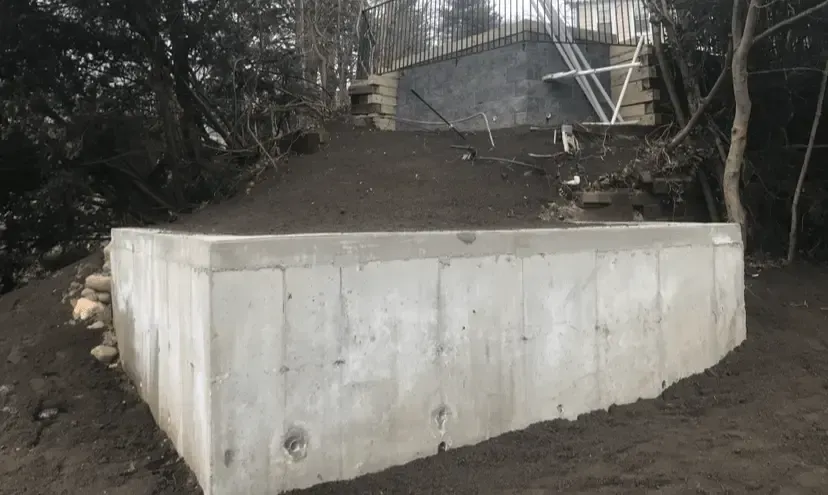 Concrete retaining walls on a hillside with stairs and trees. Gray walls, dirt and dark green trees.