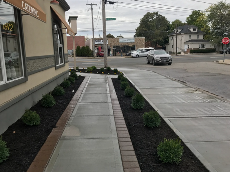 Sidewalk with bushes, brick edging, and a street corner. A car drives by on a cloudy day.