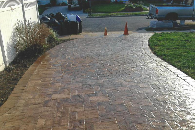 Brick-patterned driveway curves towards a street; orange cones, lawn, and a white fence are visible.