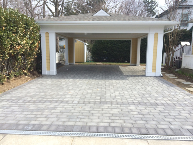Covered driveway with gray brick paving, white and yellow columns, a white roof, and a drainage system.