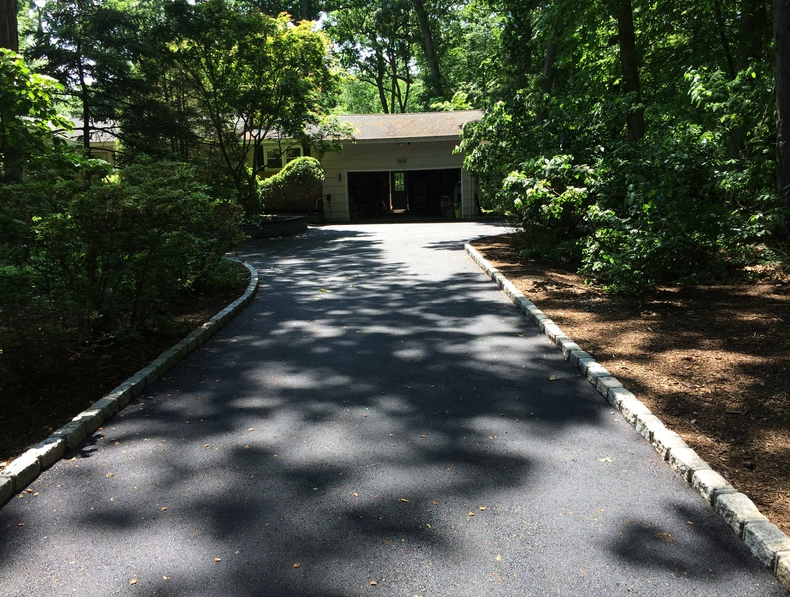 Black asphalt driveway leading to a garage, bordered by stone and surrounded by greenery.
