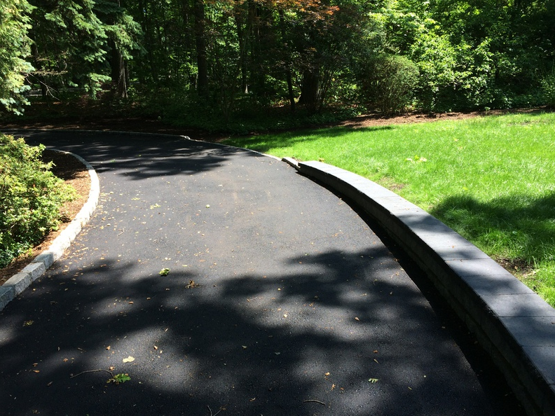 Asphalt driveway curving through a grassy yard, bordered by a low stone wall and trees.