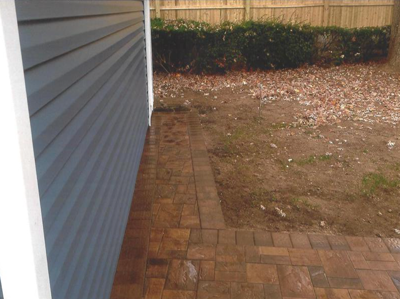 Brick patio next to a building with gray siding, leading to a grassy area with a hedge and fence.