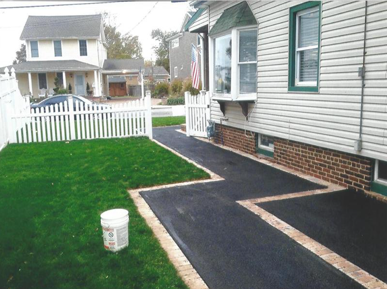 Asphalt driveway with brick border, beside a house with a white picket fence and green lawn.