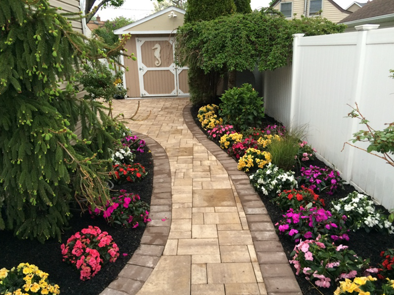 A brick pathway curves through a garden with colorful flowers and a shed.
