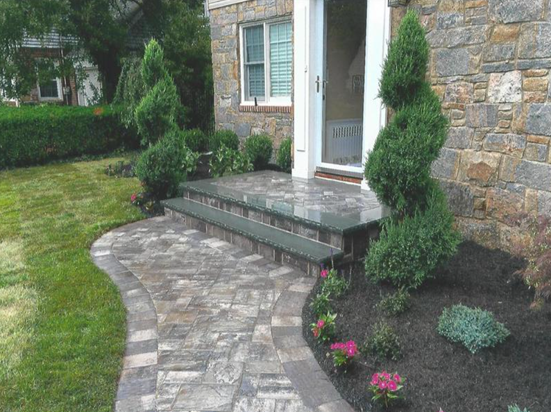 Stone walkway and steps lead to a doorway, flanked by manicured shrubs and a garden.