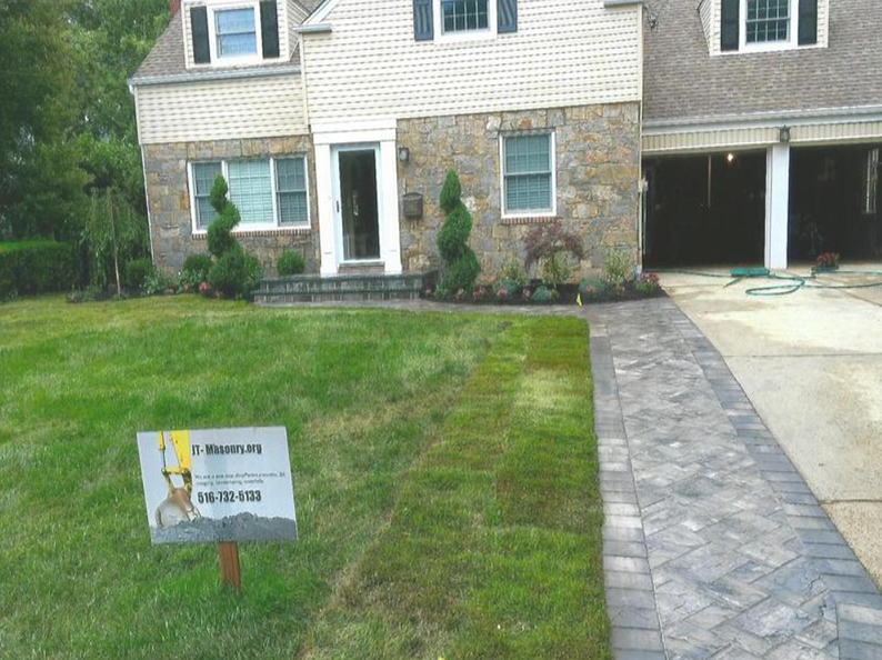House with stone facade, driveway, and landscaping. Green lawn, blue sign, and trees.