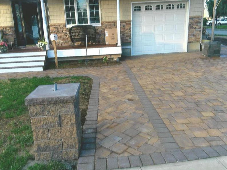 Brick paved driveway leading to a house with a garage, bordered by a low brick wall and green grass.