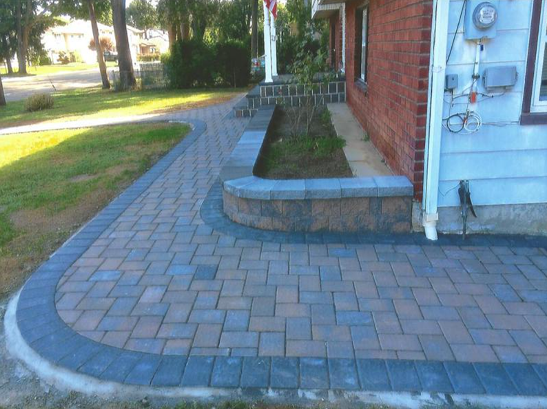 Brick walkway with a curved edge leading to a raised flower bed in front of a brick house.