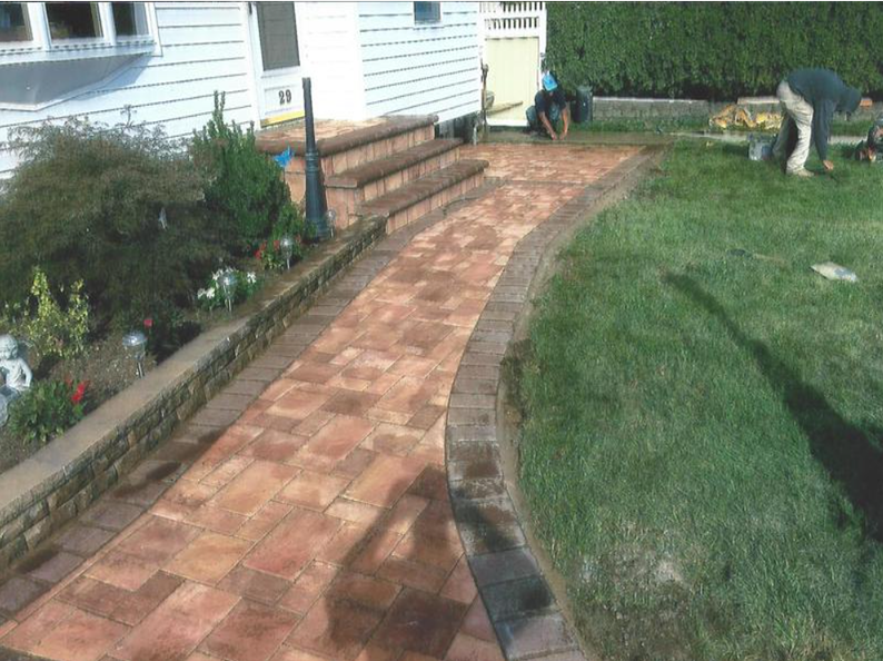 Brick pathway curves alongside lawn, leading to steps and house. Two people work in the yard.