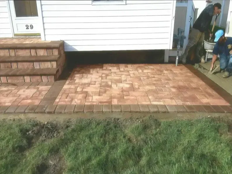 Brick patio being built near a house. Men laying pavers, next to brick steps and green grass.