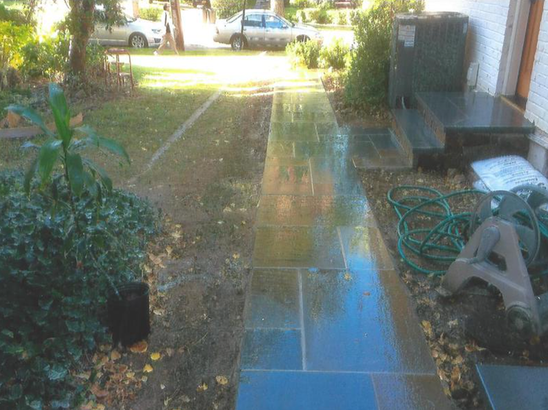 A wet, slate stone walkway leading to a house. Green grass borders on both sides.