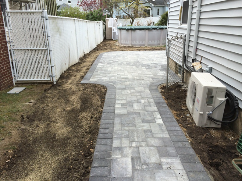 Paver pathway with gray and black borders, next to a house and fence.