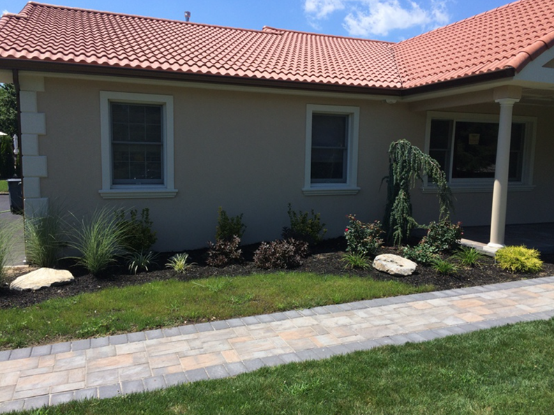 A beige house with red-tile roof, landscaped yard, pavers, and green grass.