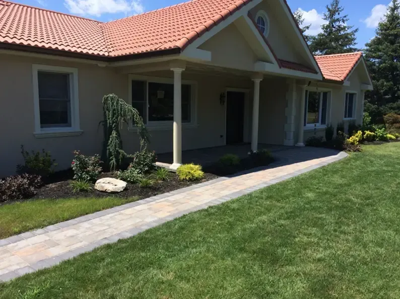 Beige house with red tile roof, porch, and paved walkway with manicured lawn.