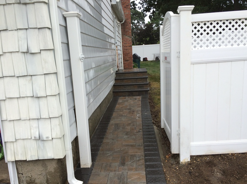 Pathway between house (left), fence (right). Brown pavers, dark brick border, steps to door.