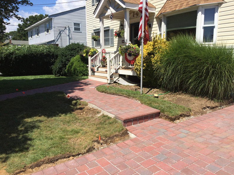 Brick pathway leading to a yellow house with white trim, American flag, and overgrown bushes.