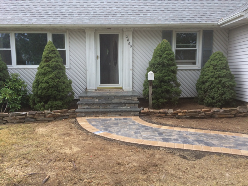 A house with stone steps and a path leading to the front door, with green shrubbery and tan and brown pavers.