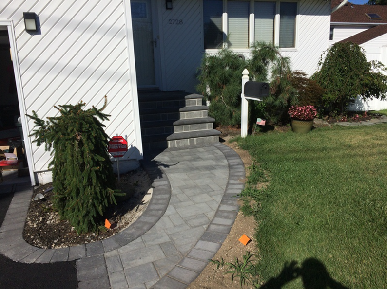A gray brick pathway and steps lead to a house entrance with a small green tree and lawn in front.