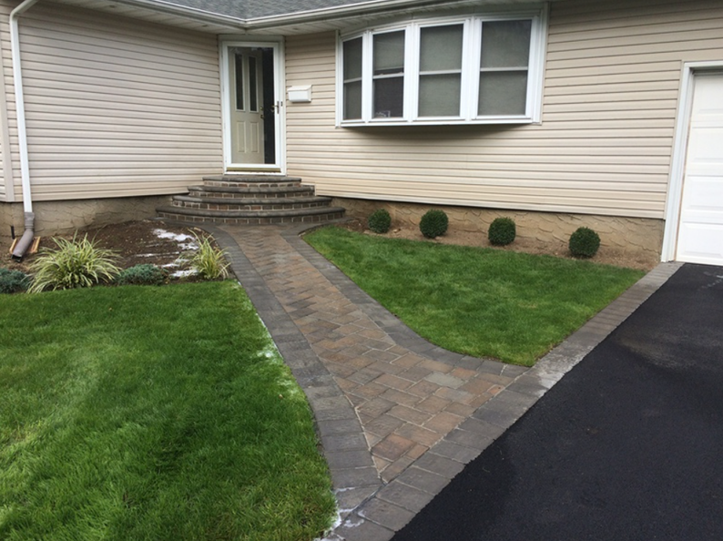 A brick path leads to a home's front door, with steps. Green grass and shrubs flank the walkway.