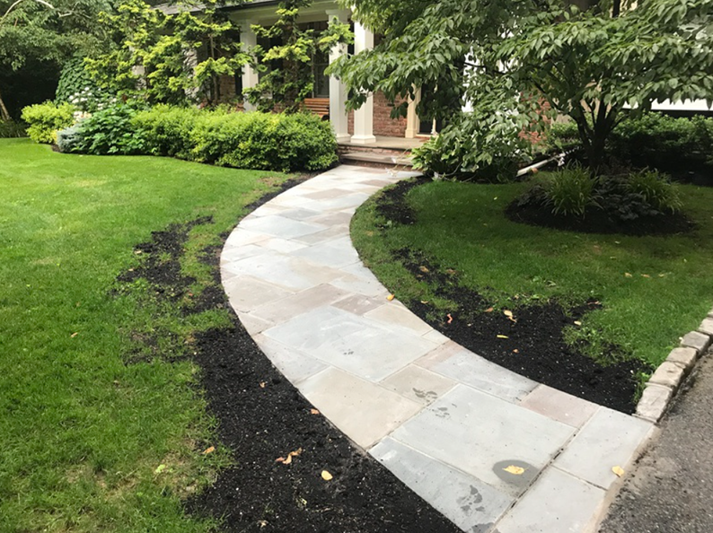 Stone walkway curving toward a house with columns; surrounded by grass and black mulch.