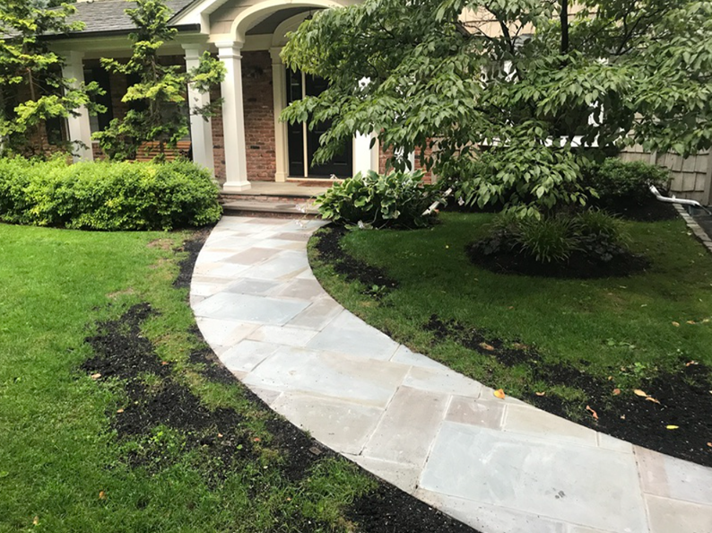 Stone walkway curves towards a house with a porch. Green grass, trees, and shrubbery surround it.