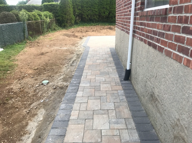 Brick pathway next to a brick and stucco building, dirt on one side, grass and fence in the background.