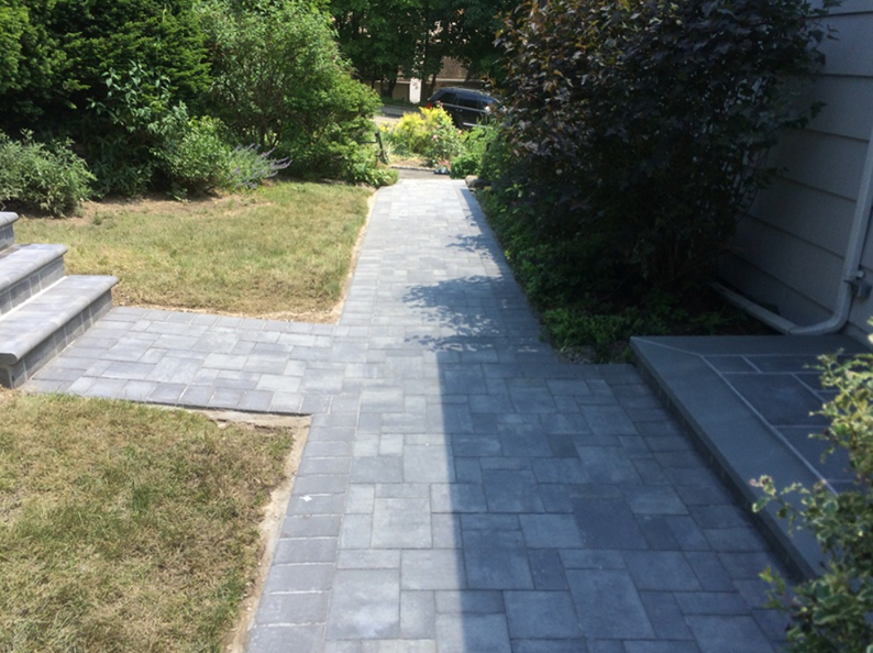 Gray paver walkway leading to a house, surrounded by grass and greenery.