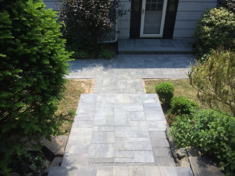 A gray stone walkway leads to a house with a dark front door, framed by green bushes and grass.