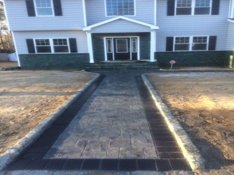 Concrete walkway leading to a house.  Gray stamped concrete with black brick border. Gray house, blue-green foundation.