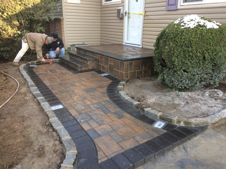 Two people installing brick walkway and steps leading to a house entrance.